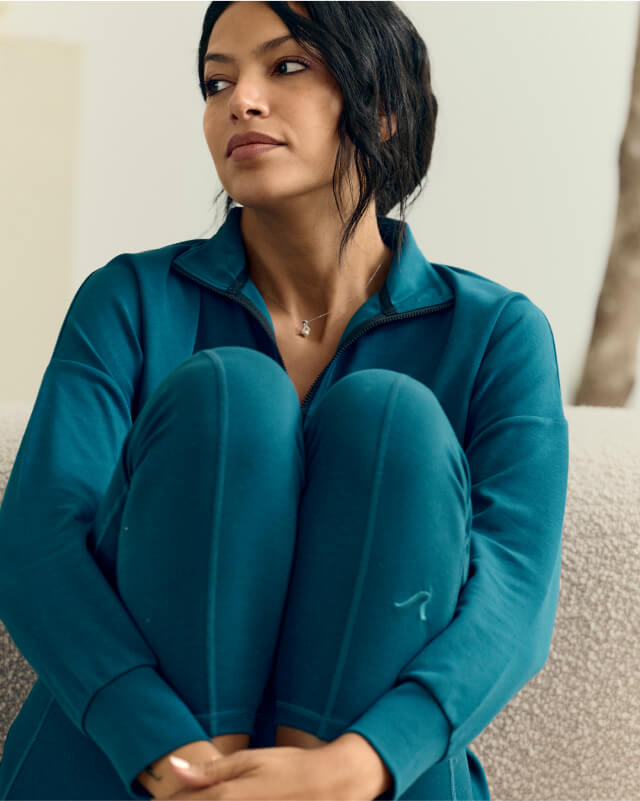 A person with dark hair sits on a light-colored couch, wearing a teal zip-up top and matching pants. They are resting their arms on their knees and looking to the side thoughtfully.
