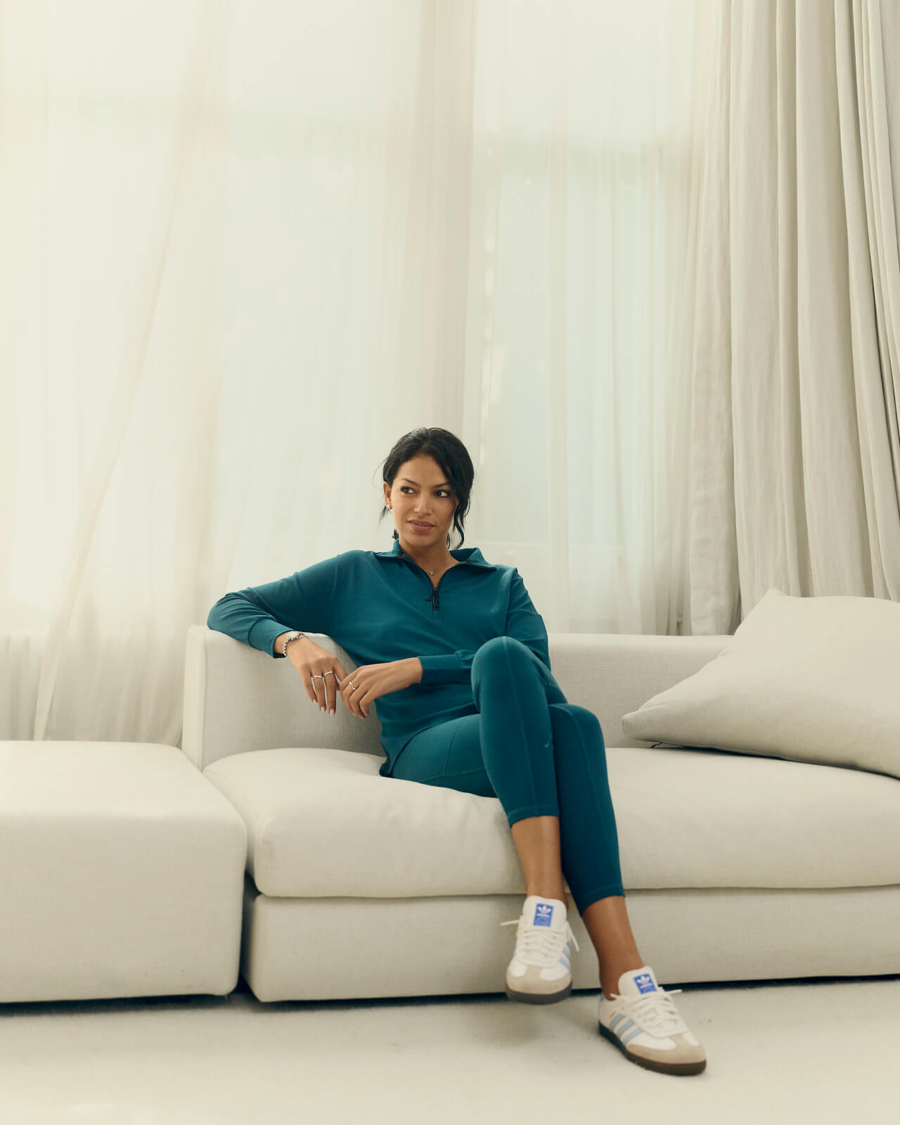 Woman in a teal tracksuit and white sneakers sits on a light-colored sofa in a softly lit room with sheer curtains. She has an relaxed expression and her hair is tied back.