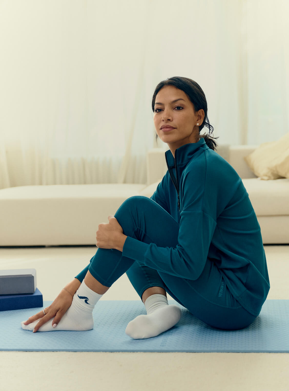 A person sitting cross-legged on a blue yoga mat indoors. They are wearing a teal tracksuit and white socks. Exercise blocks are nearby. The background features a bright, softly lit room with cream-colored furniture.