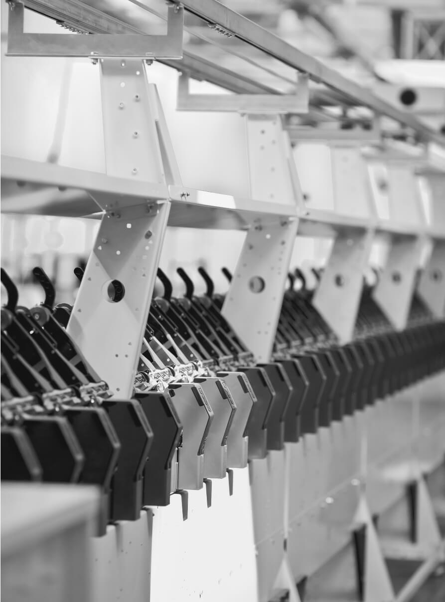 A black and white image of an industrial laundry conveyor system with multiple black metal hooks attached to a moving rail. The setup appears to be part of a larger assembly or manufacturing line.
