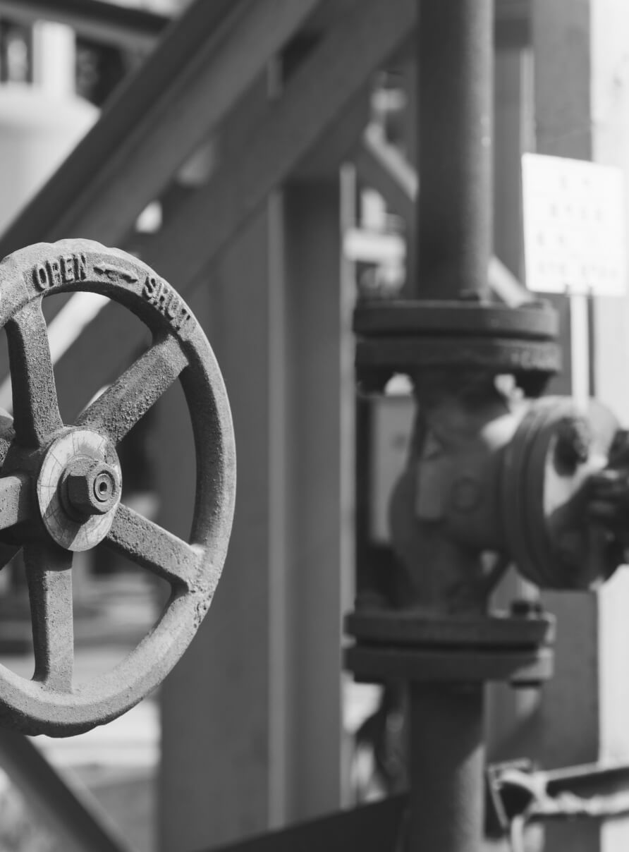 Black and white image of an industrial setting featuring a metal valve wheel with OPEN engraved on it. The wheel is attached to vertical piping, with blurred structural elements in the background.