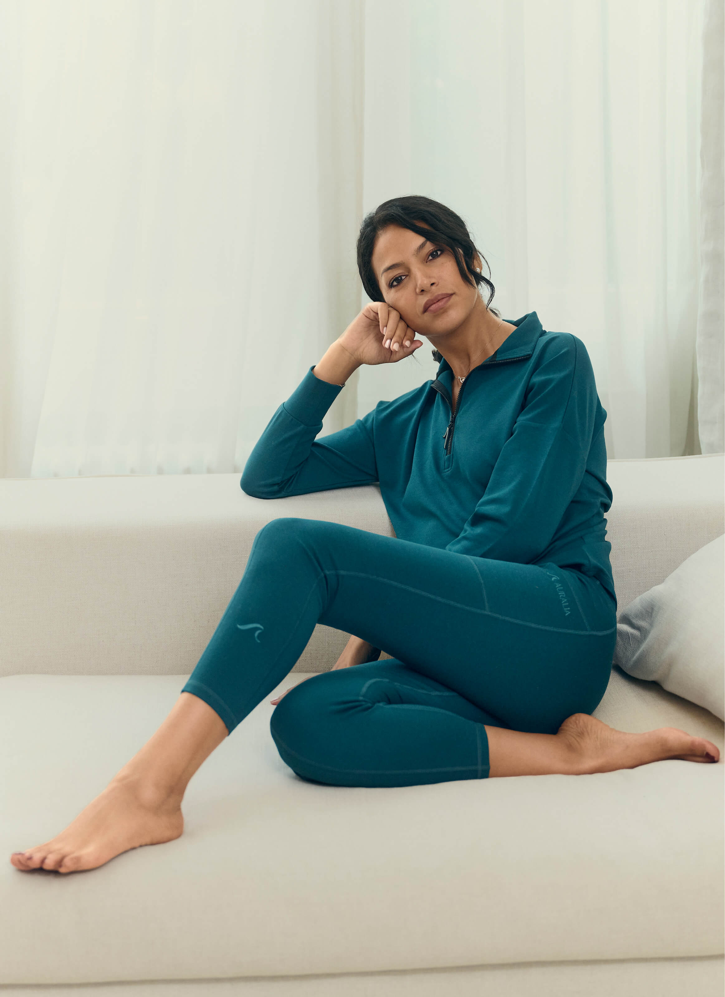 A dark-haired person sits barefoot on a light couch, wearing a teal zip-up and The Disappear Leggings from My Store, powered by AuraCloud™. Their left elbow rests on the couch back while their chin is propped on their hand. Curtains hang in the background.