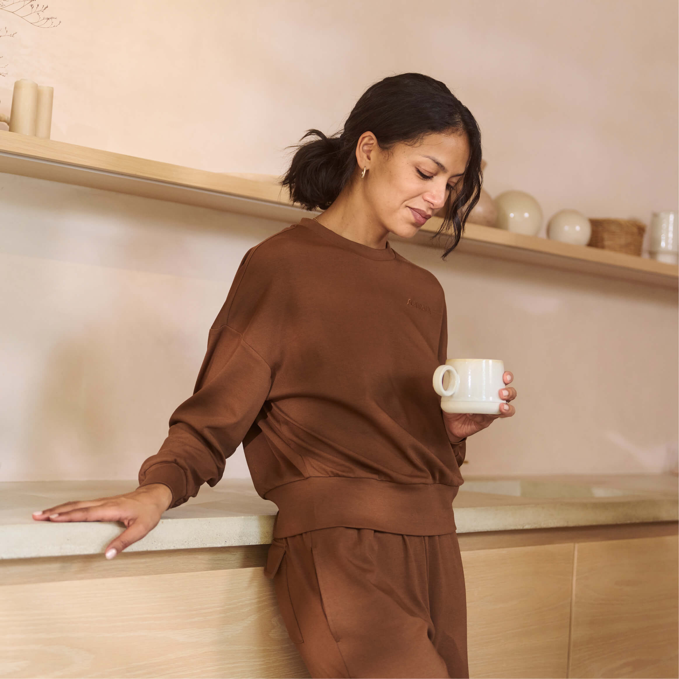 A person wearing a brown outfit stands casually in a modern kitchen, holding a white mug. Neutral-toned shelves with ceramics and a minimalist design are in the background.