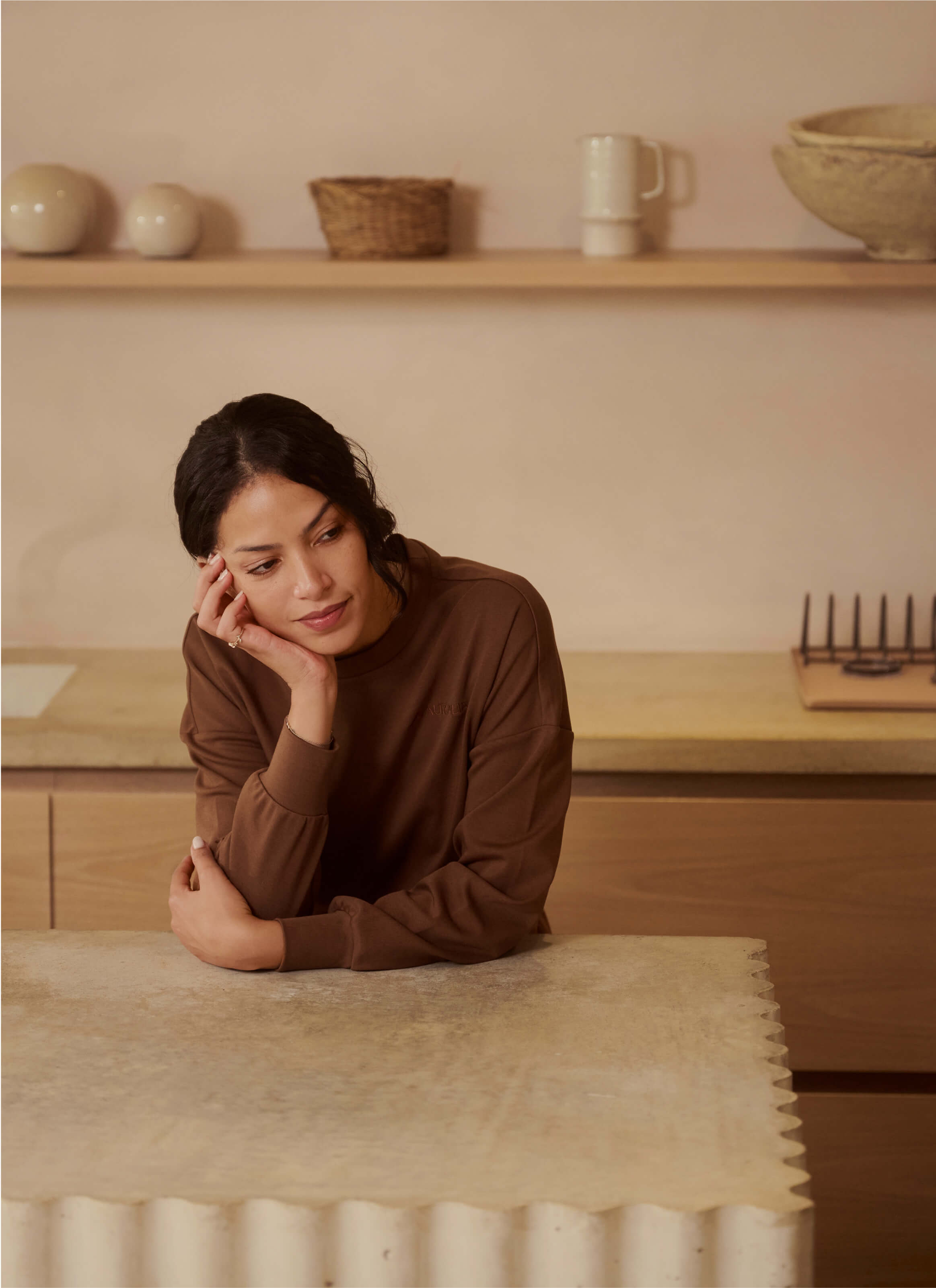 A person with dark hair leans on a textured stone counter, gazing thoughtfully while wearing My Stores Sanctuary Haven Sweatshirt in brown—a luxury long-sleeve. A wooden shelf with decorative items against a neutral-tone wall enhances the scene’s elegance.