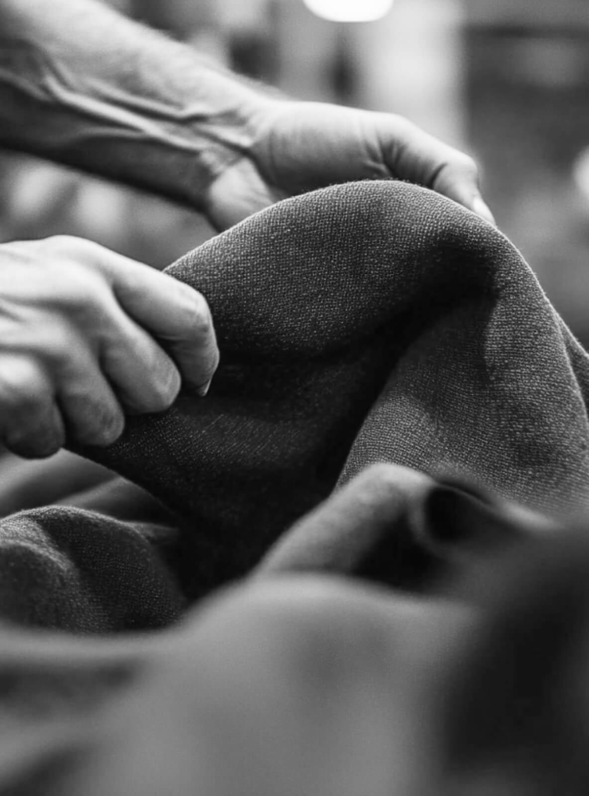 Close-up black and white image of hands holding and feeling textured fabric. The focus is on the interaction between the hands and the material, highlighting the fabrics weave and movement.