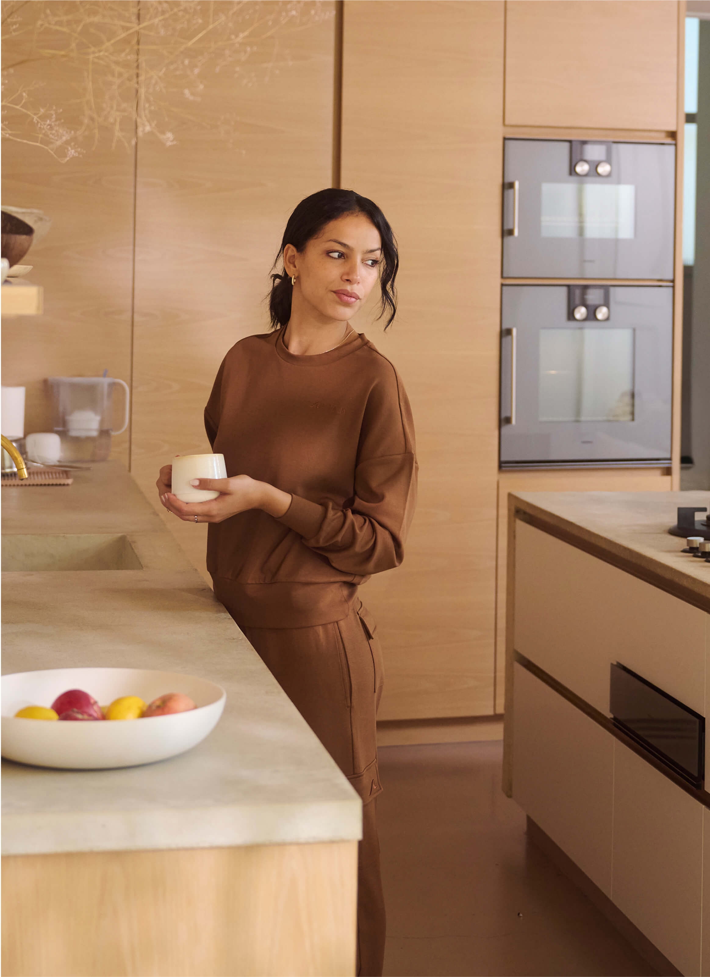 A woman in The Sanctuary Cocoon Joggers by My Store holds a mug in a modern kitchen with light wood cabinets and built-in ovens. She gazes thoughtfully to the side, while a bowl of fruit adds vibrancy to the sustainable space.