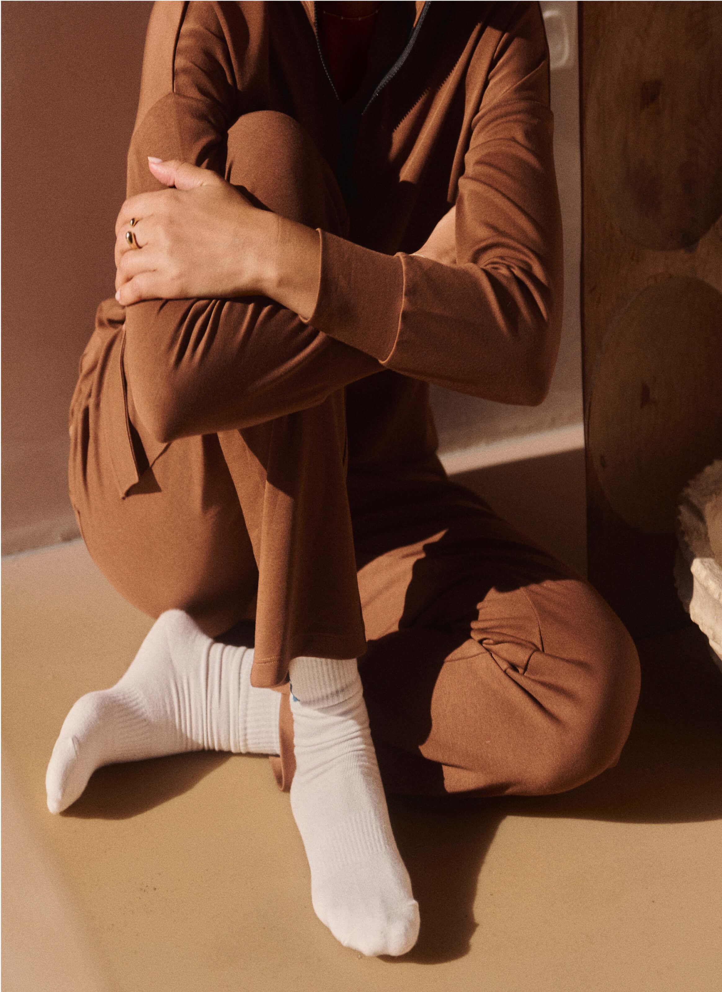 In a warmly lit room with cut wood in the background, someone sits comfortably on the floor wearing The Sanctuary Cocoon Joggers from My Store. Dressed in brown and cozy white socks, they wrap their arms around their knees, possibly contemplating sustainable choices.