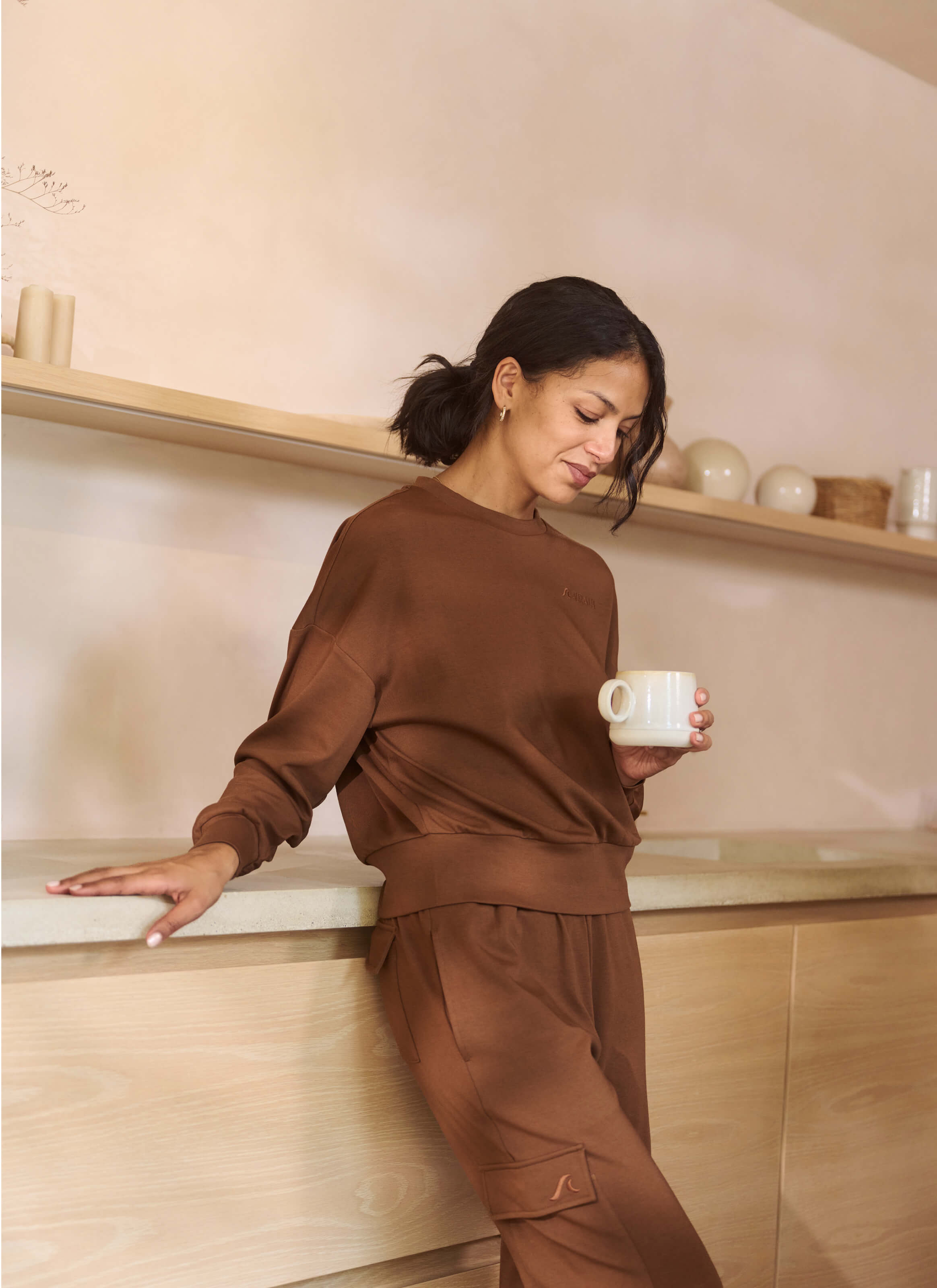 A person wearing The Sanctuary Cocoon Joggers from My Store leans against a kitchen counter with a white mug, slightly smiling. Minimalist shelves with spherical decor and a basket add to the cozy atmosphere.