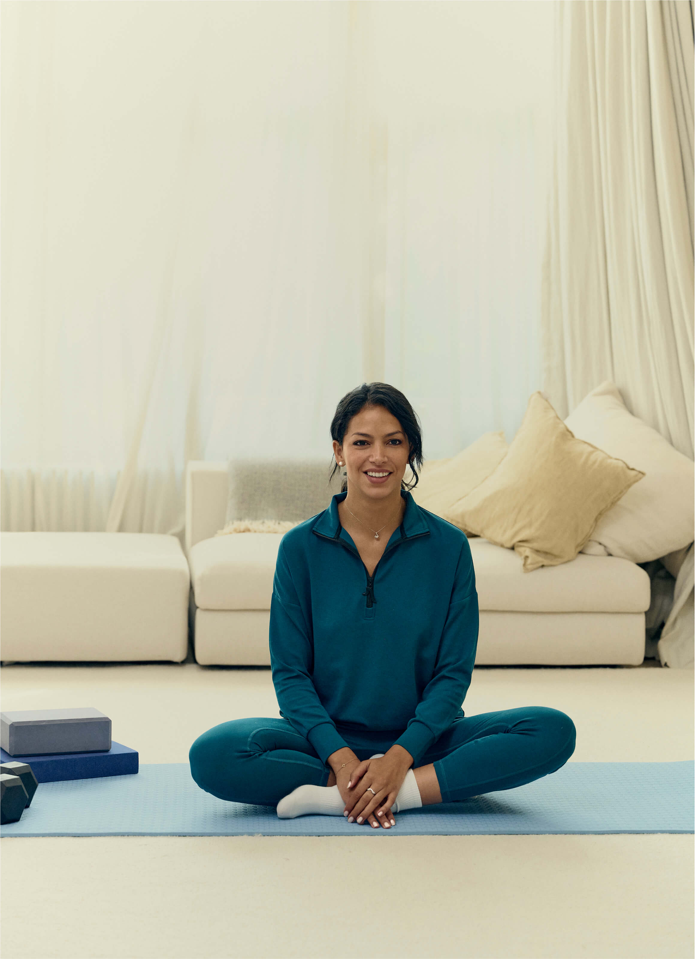 A person sits cross-legged on a yoga mat in The Sanctuary Quarter-Zip Pullover in turquoise from My Store, wearing white socks. Beige and white pillows adorn the sofa behind them, enhancing serenity. Fitness blocks rest at the mats edge, completing this sustainably sourced peaceful scene.