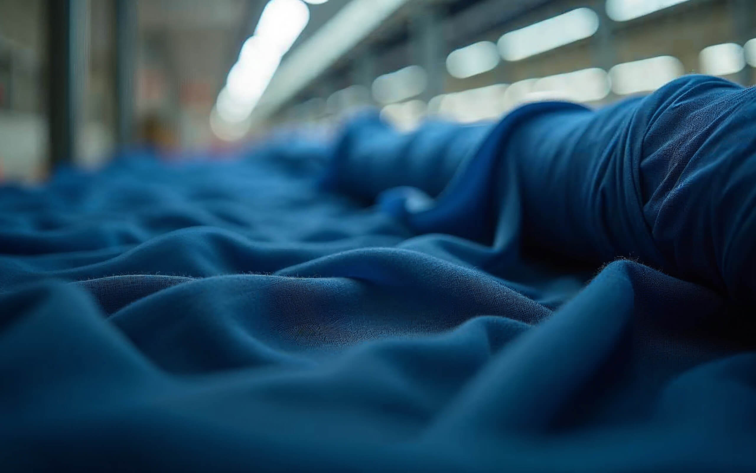 Rolls of soft, blue fabric are spread out and layered on a flat surface in a warehouse or manufacturing setting. The fabric is slightly rumpled, with a blurred, well-lit background indicating an industrial space.