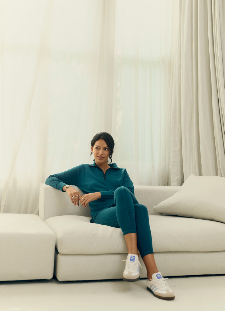 Woman in teal loungewear sits casually on a white sofa in a bright room with sheer curtains. She has her legs crossed and wears white sneakers. The atmosphere feels calm and relaxed.