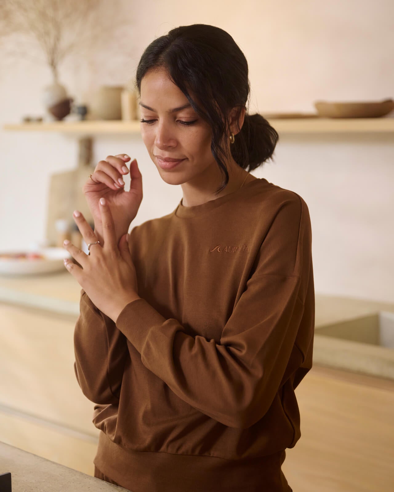 Woman with dark hair tied back, wearing a brown long-sleeve shirt, stands in a softly lit room with minimalistic decor. She looks down while gently touching her fingers. Shelves with vases and neutral tones in the background.