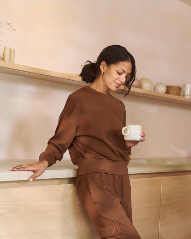 A person in a brown outfit leans against a light wood counter, holding a white mug. Behind them is a shelf with decorative items and a beige wall. The setting is minimalistic and warmly lit.