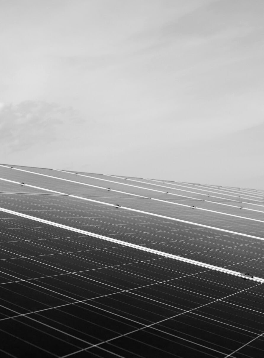 Monochrome image of an array of solar panels angled upwards, set against a cloudy sky. The panels have a grid-like pattern and occupy the lower portion of the image, with the sky taking up the upper part.