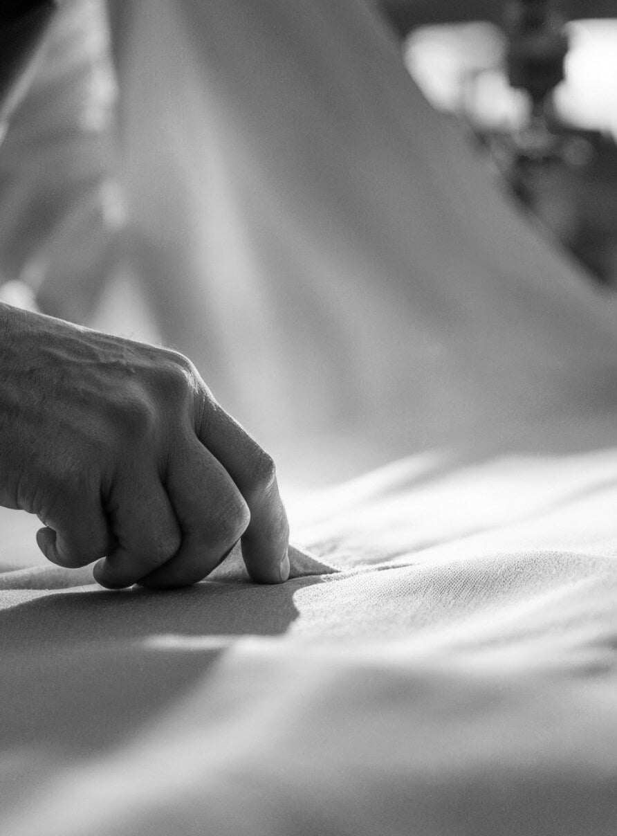 Close-up of a hand gently running across a smooth fabric under bright light, casting soft shadows. The black and white image highlights the texture of the fabric and the delicacy of the touch.