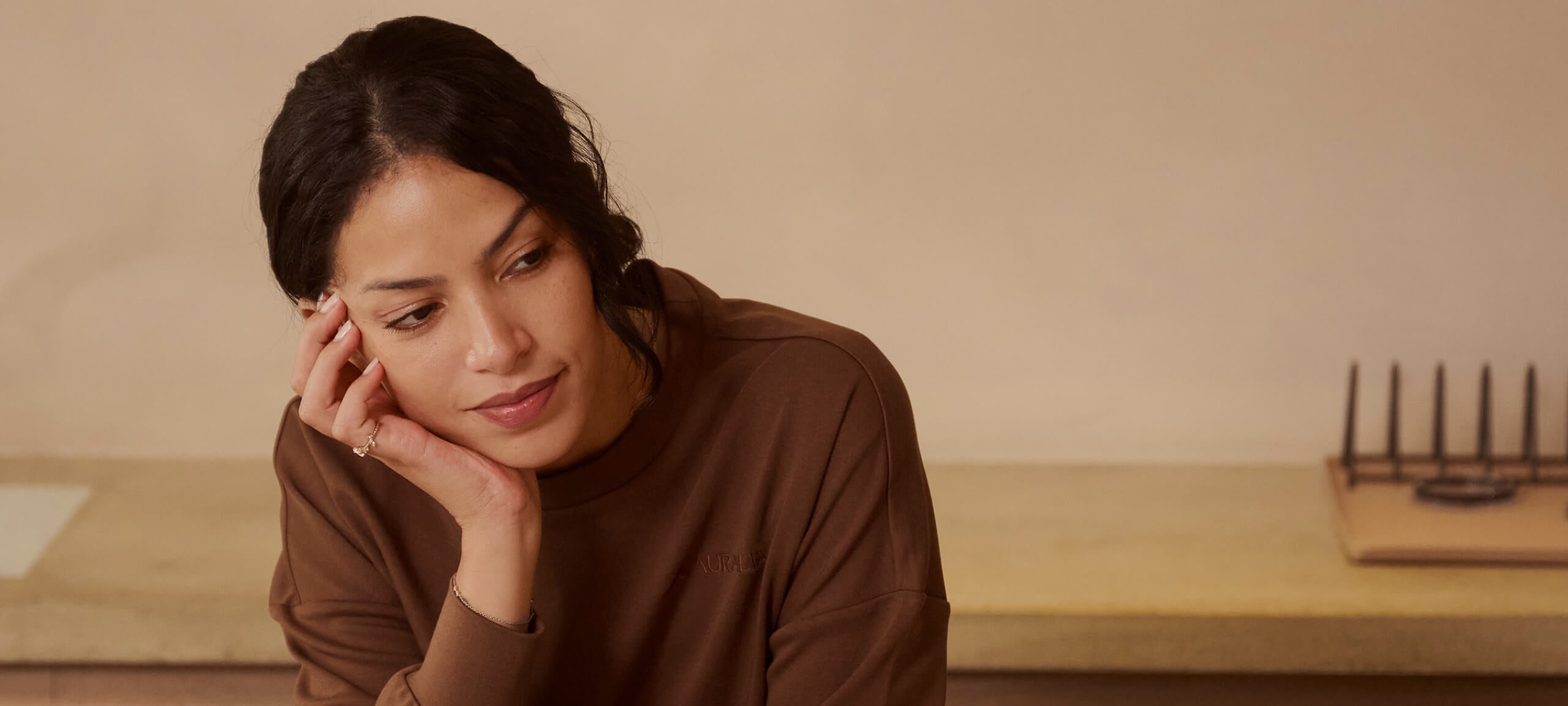 A person with dark hair is sitting with their hand resting on their cheek, looking thoughtfully to the side. They are wearing a brown sweater. The background is neutral and blurred.