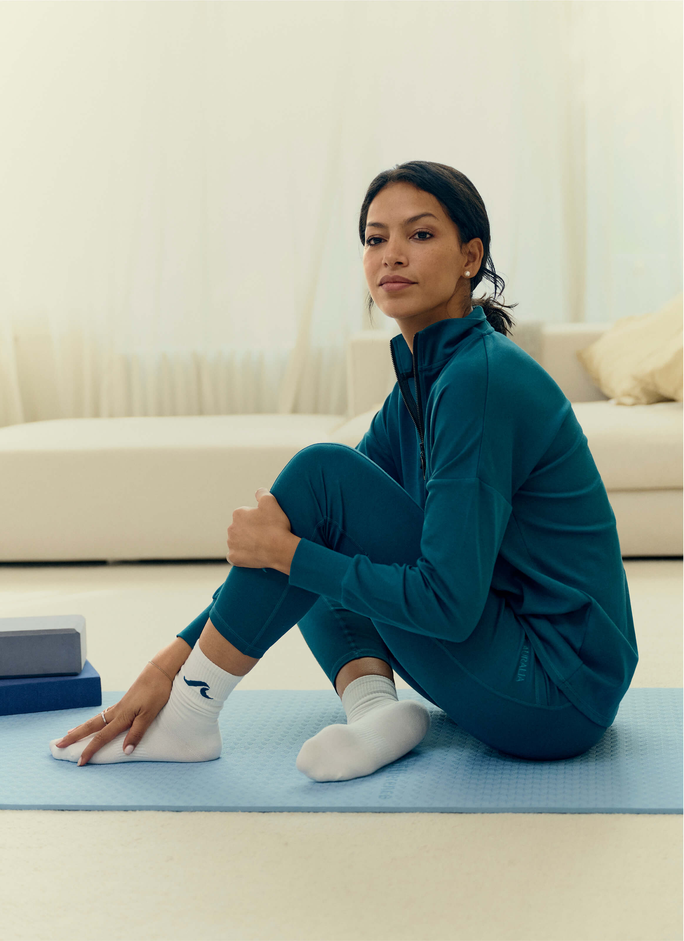 A person sits cross-legged on a blue yoga mat, wearing The Disappear Leggings by My Store in teal modal fabric. White socks complete the outfit as they gaze calmly ahead. Yoga blocks and an AuraCloud™ vibe enhance comfort under soft light near the beige sofa.