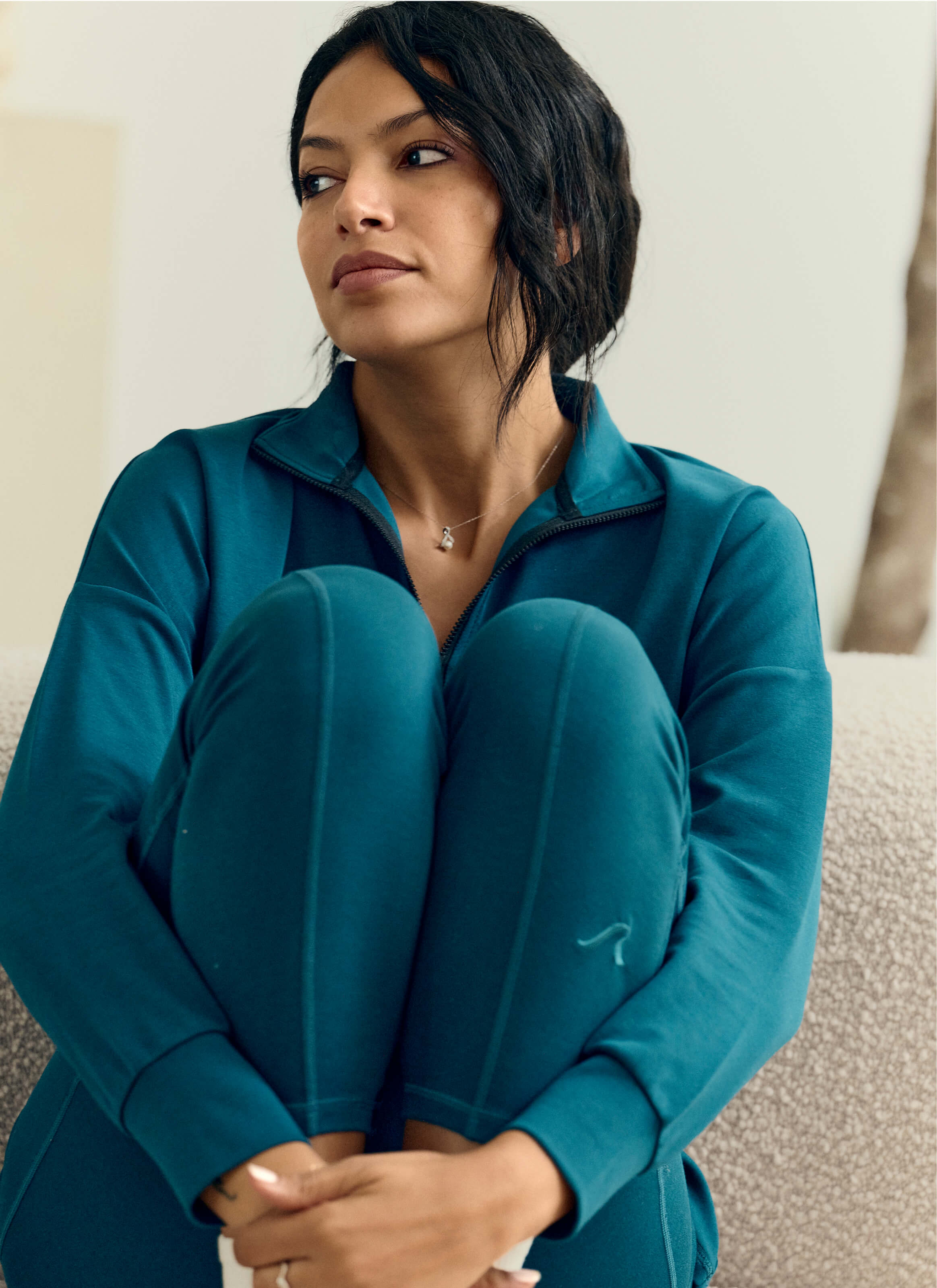 A person with long dark hair sits with knees up, wearing a teal zip-up jacket and The Disappear Leggings - Powered by AuraCloud™ by My Store, made from sustainable modal. They look thoughtfully to the side against a soft-focus indoor backdrop.
