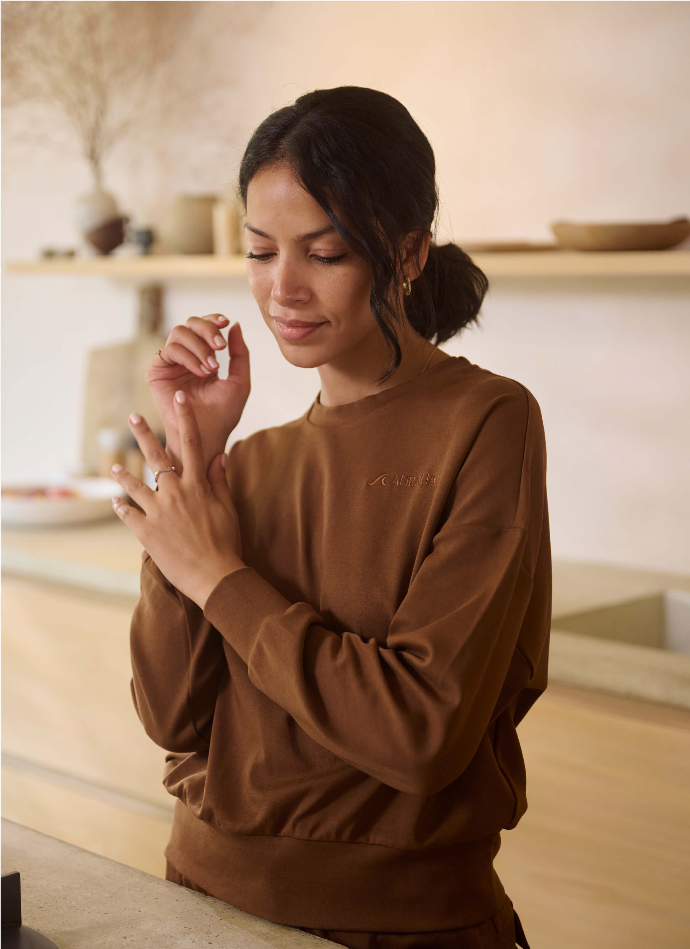 A person with dark hair tied back stands in a softly lit room, gently rubbing their hands. They wear The Sanctuary Haven Sweatshirt from My Store, a relaxed-fit, brown luxury fabric top, and smile lightly. Shelves with neutral-colored decor adorn the background.