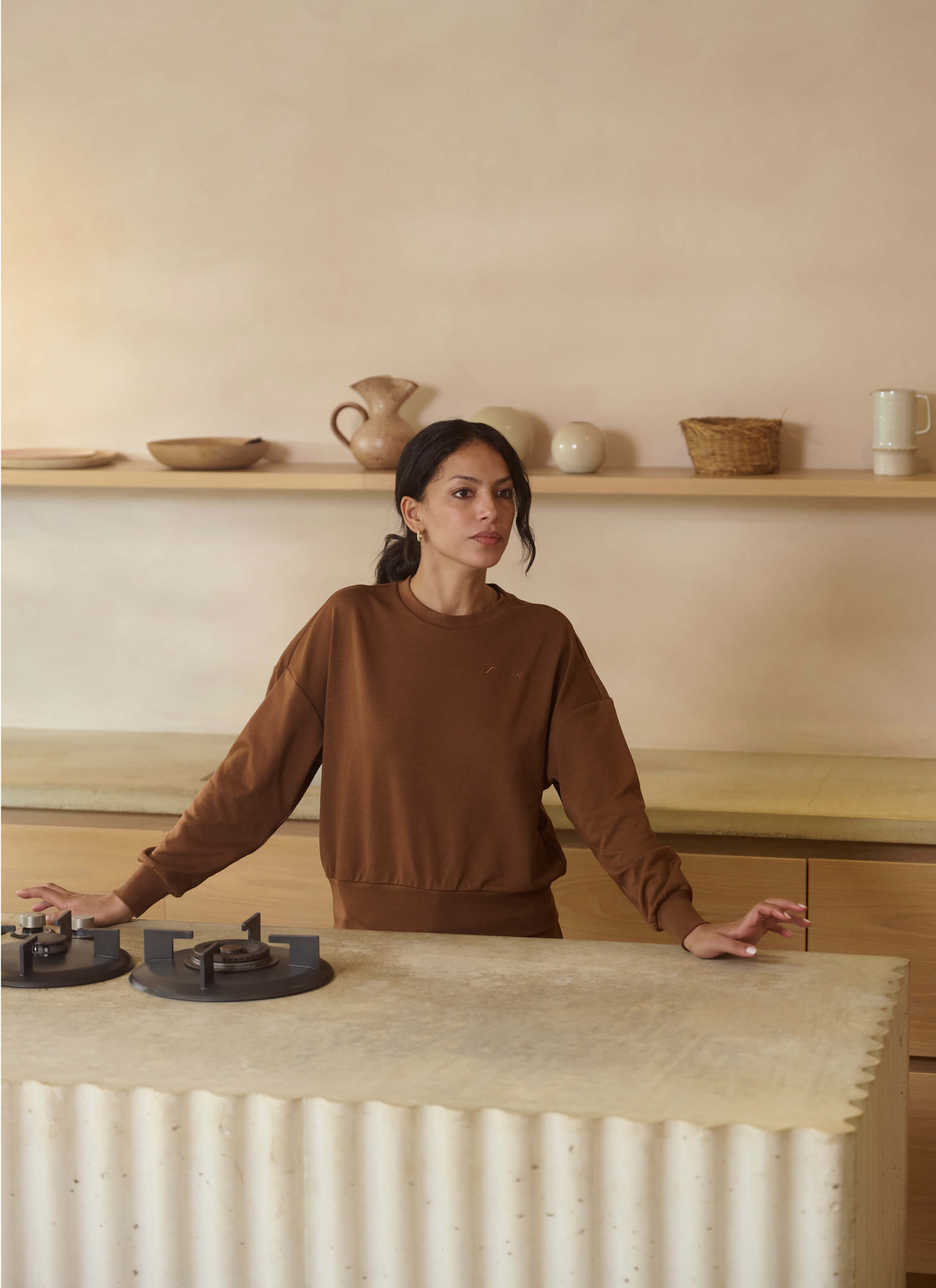A person in a relaxed fit, Sanctuary Haven Sweatshirt by My Store stands in a minimalist kitchen. They lean on a marble counter with a stovetop. Wooden shelves display bowls, vases, and baskets. Light beige walls enhance the sophisticated ambiance.