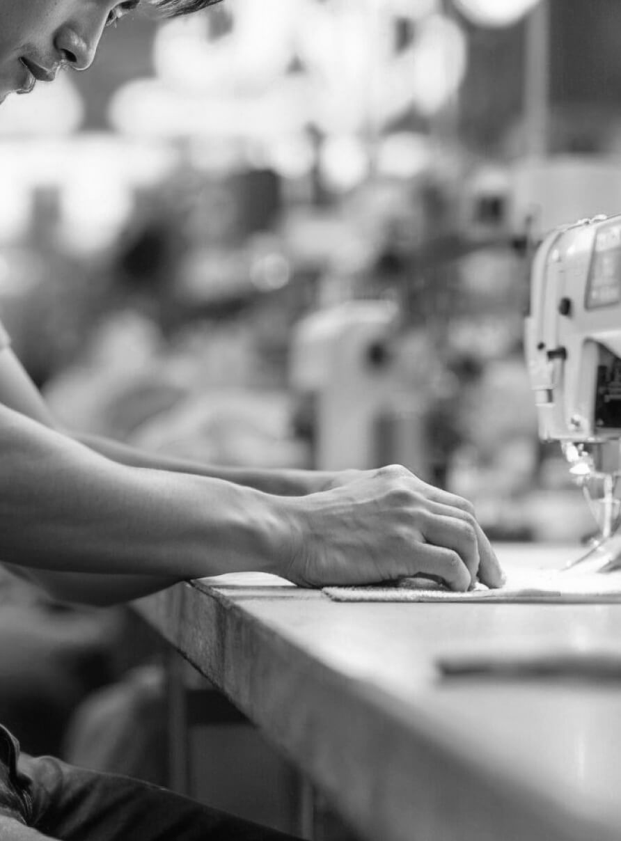 Black and white image of a person sewing fabric on a sewing machine. The focus is on the hands guiding the cloth, while blurred sewing equipment is visible in the background.