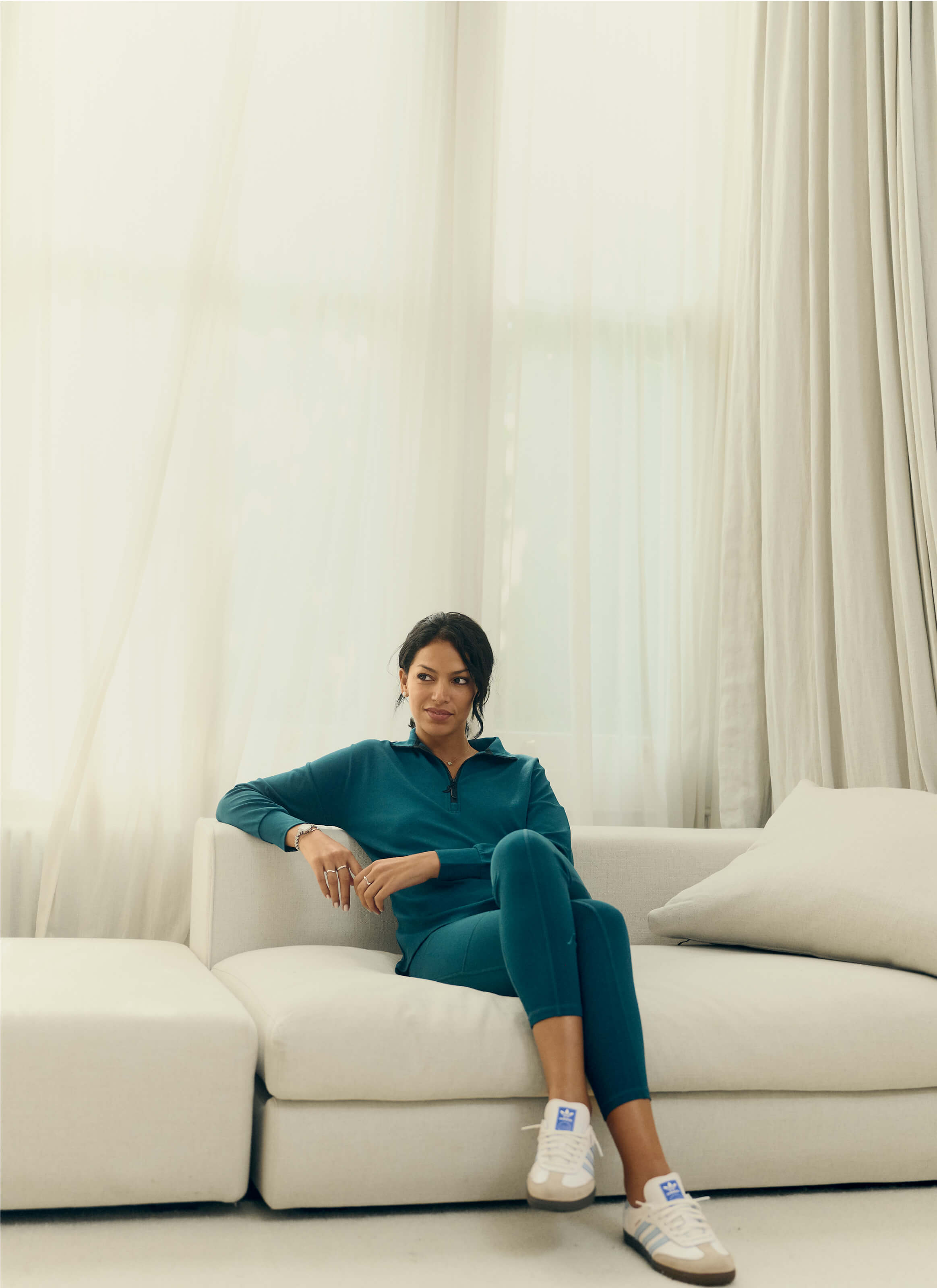 A person sits on a white sofa in a softly lit room with sheer curtains, donning the eco-elegant Sanctuary Quarter-Zip Pullover by My Store, powered by AuraCloud™, complemented with white sneakers. The sustainably sourced ensemble enhances the serene and relaxed atmosphere.