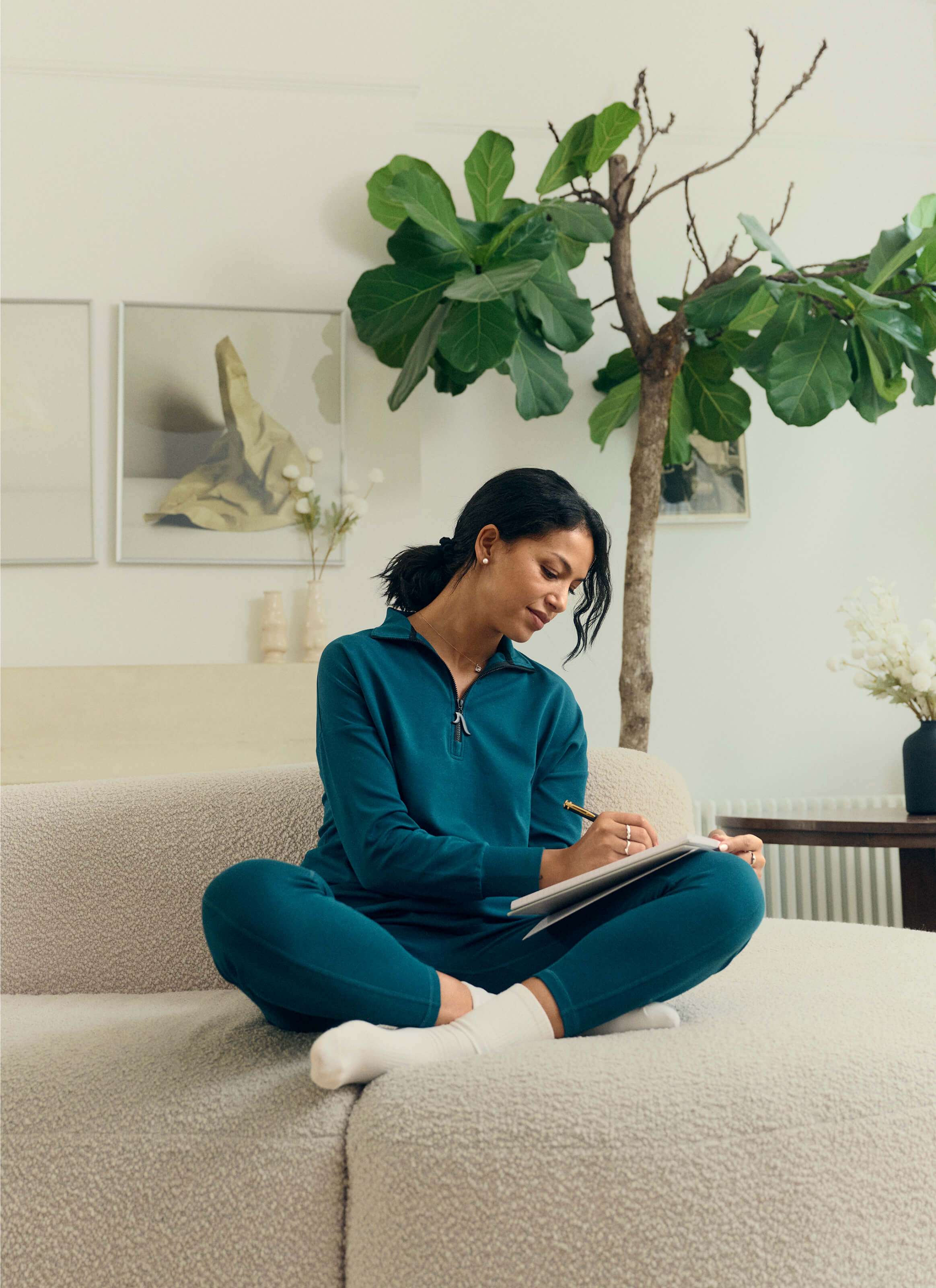 Dressed in The Sanctuary Quarter-Zip Pullover from My Store, a person sits cross-legged on a beige sofa, jotting down notes. A large plant and framed artworks enhance the rooms eco-elegant comfort, exuding AuraCloud™ sustainability for a calming and cozy atmosphere.