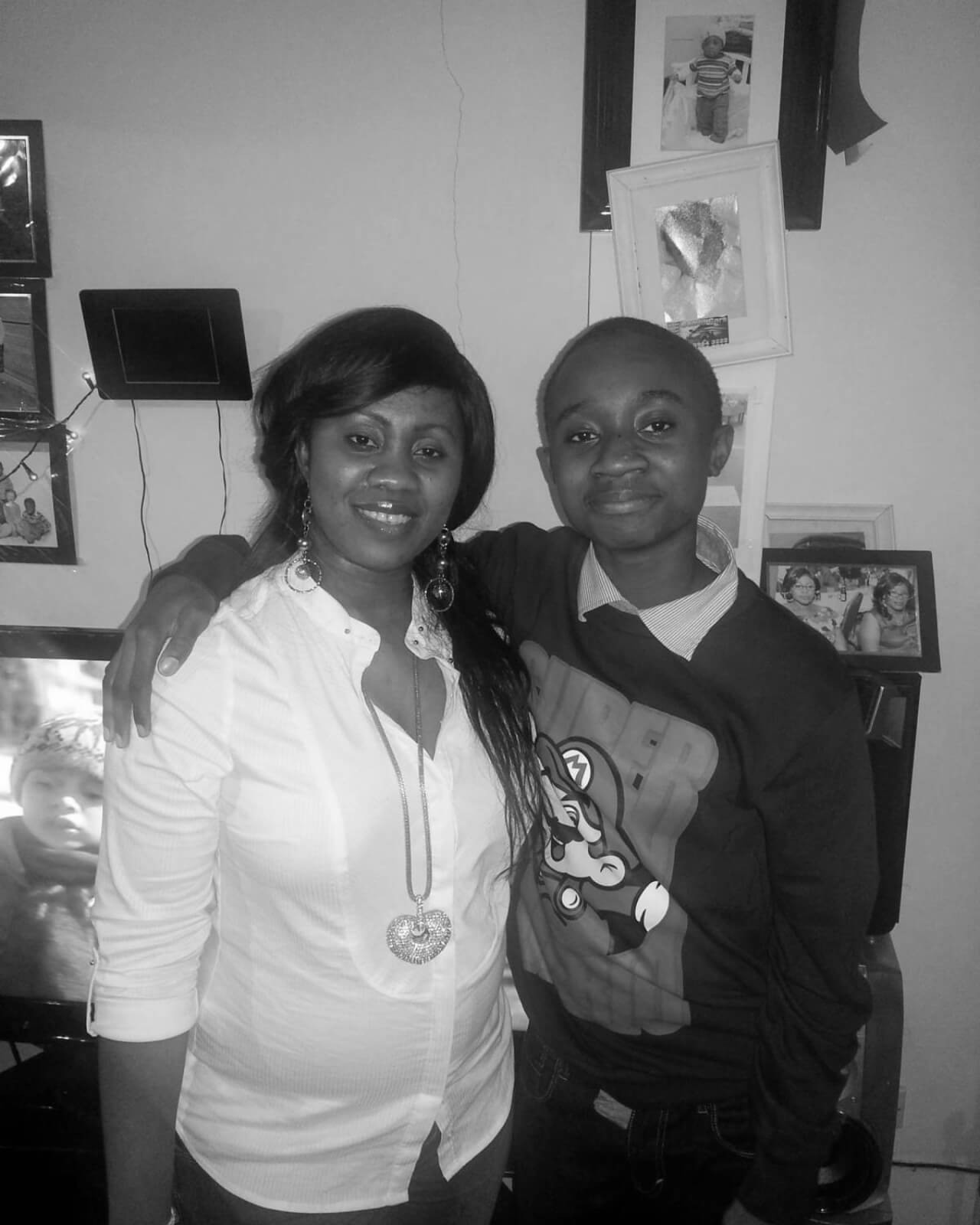 A black and white photo of two people standing close together indoors. The person on the left wears a white blouse and necklace, while the person on the right wears a sweater with a design. They are both smiling. Various framed photos are visible in the background.