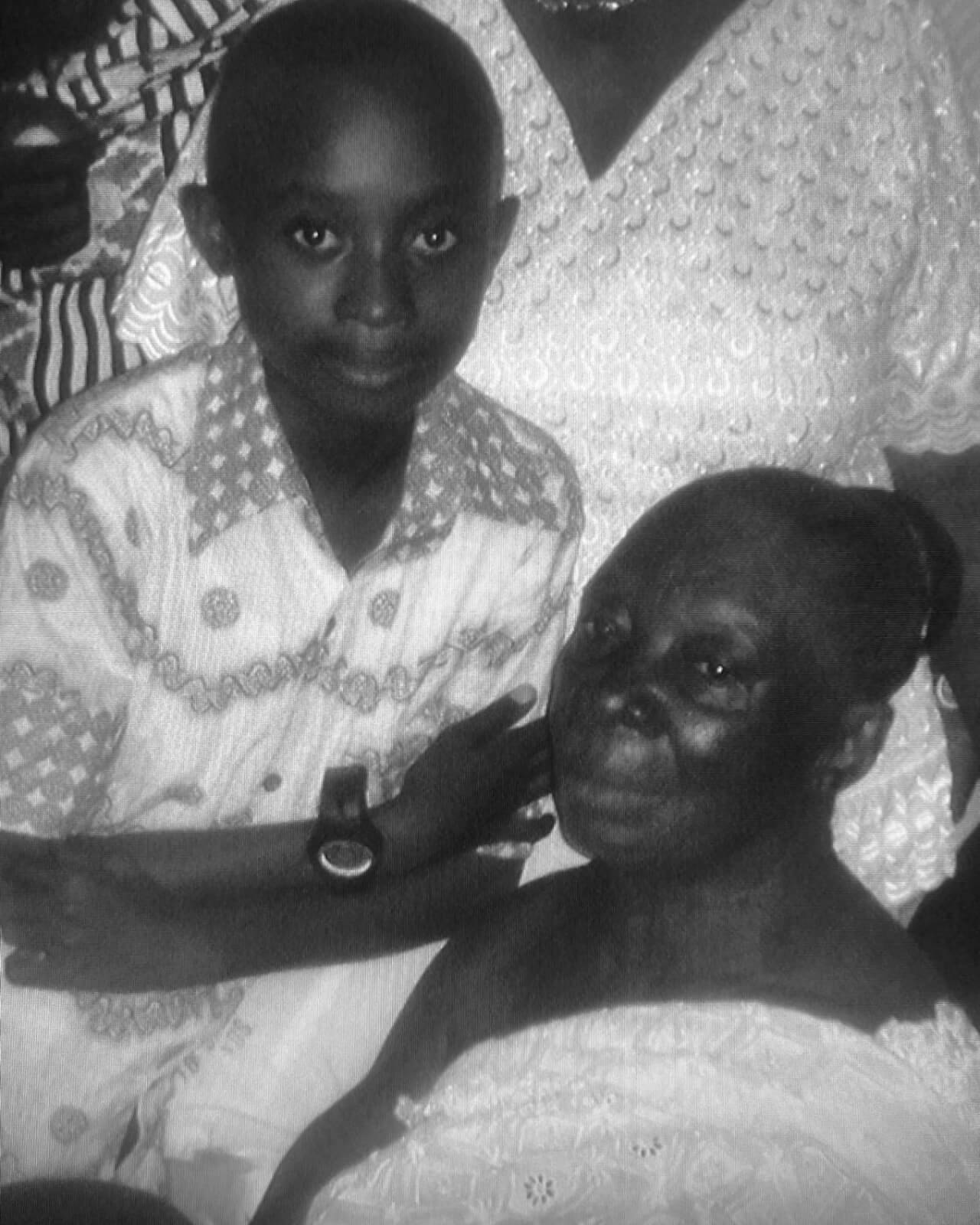 A young boy in a patterned shirt stands beside an elderly woman seated in a patterned dress. They both look toward the camera with gentle expressions. The photo is in black and white, with others partially visible in the background.