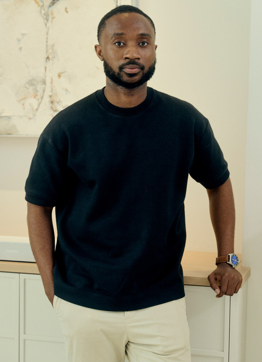 A man with short hair and a beard is leaning against a light-colored piece of furniture. He is wearing a black t-shirt, beige pants, and a watch. The background features a light abstract painting on the wall.