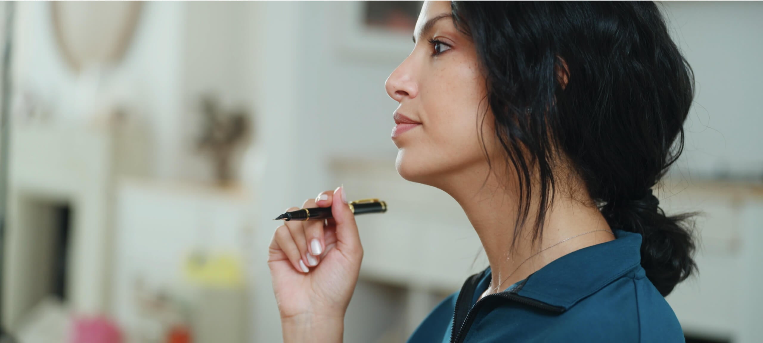 A woman with dark hair tied back holds a pen near her face, appearing contemplative. She is wearing a blue top and looking thoughtfully off to the side, set against a softly blurred background.